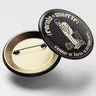 Pin tipo botón abierto con diseño esqueleto “escala a muerte”, vista del broche interior metálico.