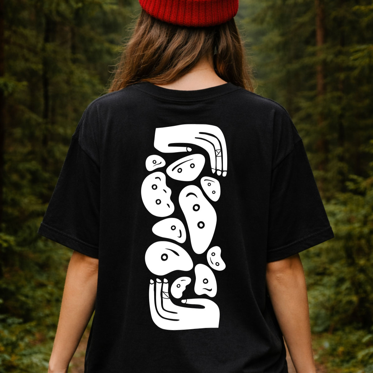Mujer de espaldas con gorro rojo usando playera Totem Holds negra con diseño de tótem de agarres de escalada en la espalda, en bosque.