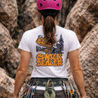 Mujer de espaldas usando playera de escalada Chalk Dealer con casco rosa en pared de roca, diseño grande en la espalda