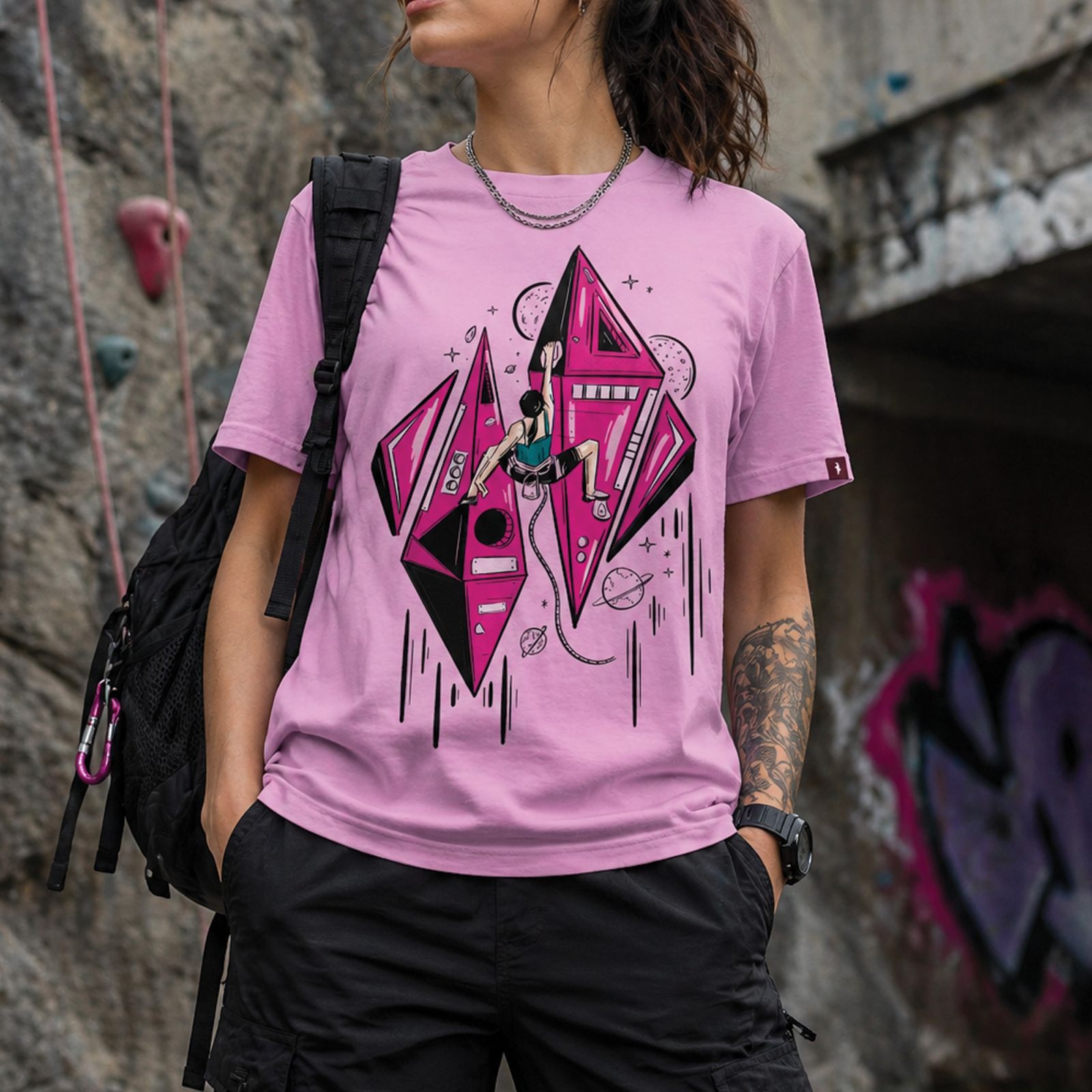 Modelo usando playera rosa lavanda Cosmic Climb con ilustración frontal de escalada espacial frente a un muro de escalada.